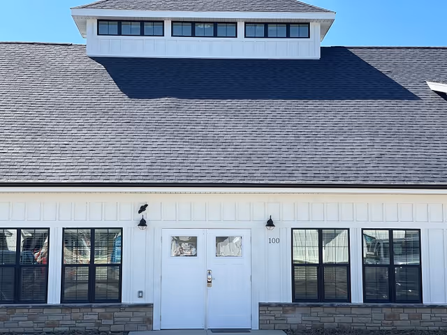 Front exterior view of a building with white siding, black-framed windows, a double white door with the number 100, and a dark shingled roof under a clear blue sky.
