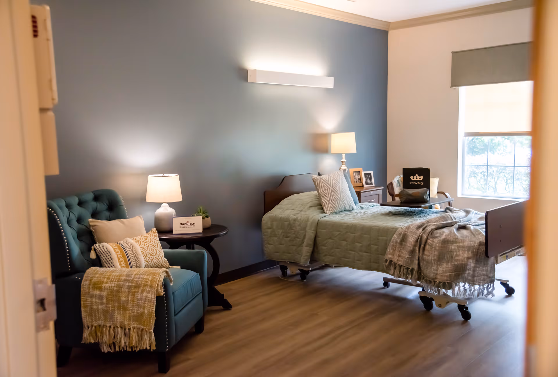 A cozy nursing facility bedroom with a single adjustable bed, bedside tables and lamps, and a teal armchair by a window.