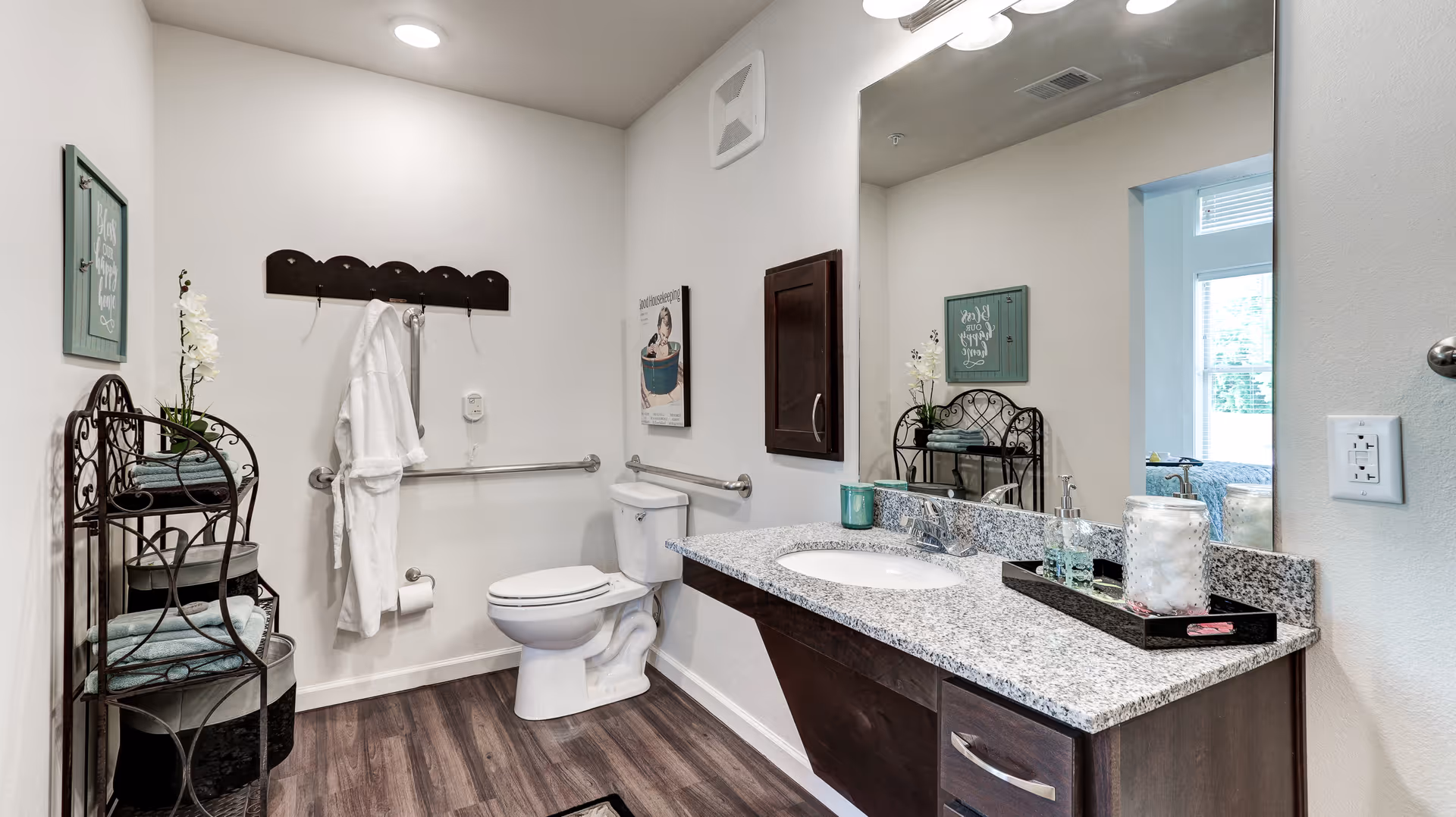A clean and modern bathroom with a white toilet, a granite countertop with a sink, and a large mirror above the sink. There is a dark wooden cabinet mounted on the wall, a towel rack with a white robe hanging, and a decorative metal shelf holding folded towels and a potted orchid. The floor is wood-style laminate, and the walls are painted white. A window allows natural light into the room.