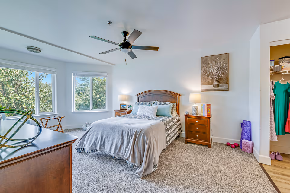 A bright and cozy bedroom with a large bed covered in gray and patterned bedding, two wooden nightstands with lamps, a ceiling fan, two large windows showing greenery outside, a dresser with decorative items, and an open closet with hanging clothes and yoga mats on the floor.