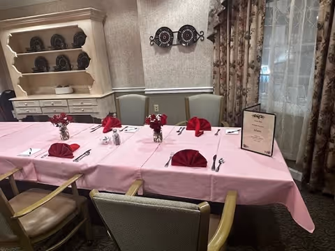 A dining room table set with a pink tablecloth, red folded napkins, silverware, and small vases with red flowers. The table is surrounded by chairs, and there is a menu placed on the table. In the background, there is a white hutch with plates and decorative items, floral curtains, and a wall with two decorative circular wall hangings.