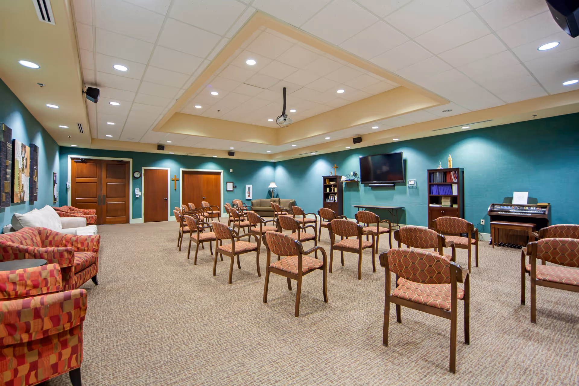 A spacious meeting or activity room with multiple rows of wooden chairs with patterned upholstery arranged facing a wall-mounted flat screen TV. The room has teal walls, beige carpet, recessed lighting, and a ceiling-mounted projector. There are two wooden bookshelves, a small table, a piano, and a few couches along the walls. Two wooden doors and a double door are visible on one side of the room.