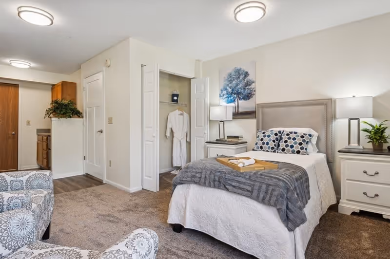 A cozy bedroom in a senior living facility featuring a neatly made bed with white and gray bedding, two patterned pillows, and a gray throw blanket. There are two white nightstands on either side of the bed, each with a lamp and decorative items. A closet with open double doors reveals a hanging white robe. The room has beige carpet, light-colored walls, and a painting of a tree above the bed. In the foreground, there are two patterned armchairs. A small kitchen area is visible in the background.