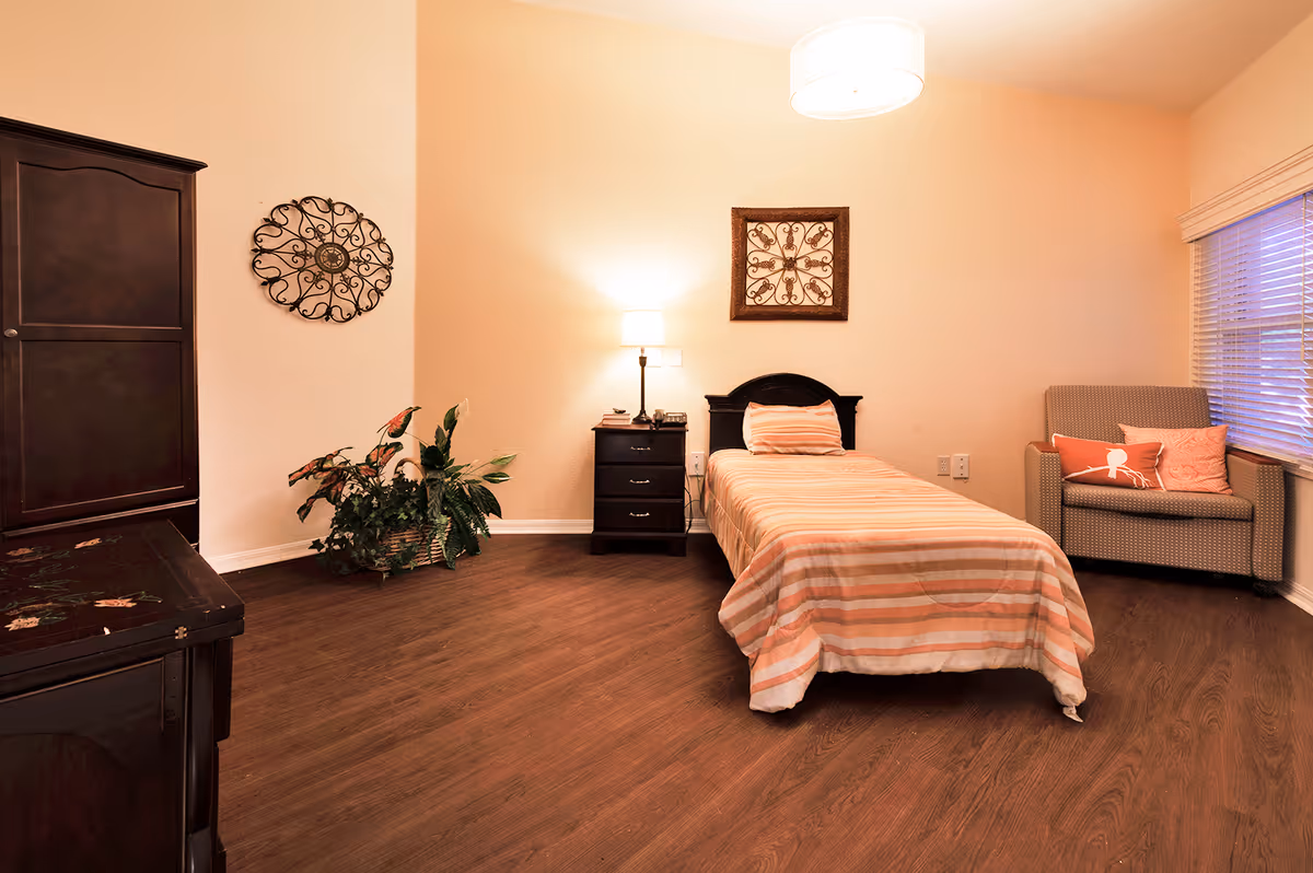 A warm, neatly furnished bedroom with a single bed, nightstand and lamp, armchair by a window, wooden flooring and decorative wall art.