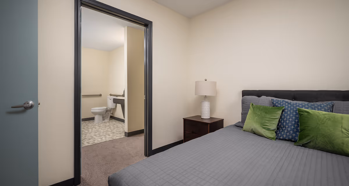 Bedroom with a neatly made bed, nightstand and lamp, and an open doorway showing an adjacent bathroom with a toilet and sink.