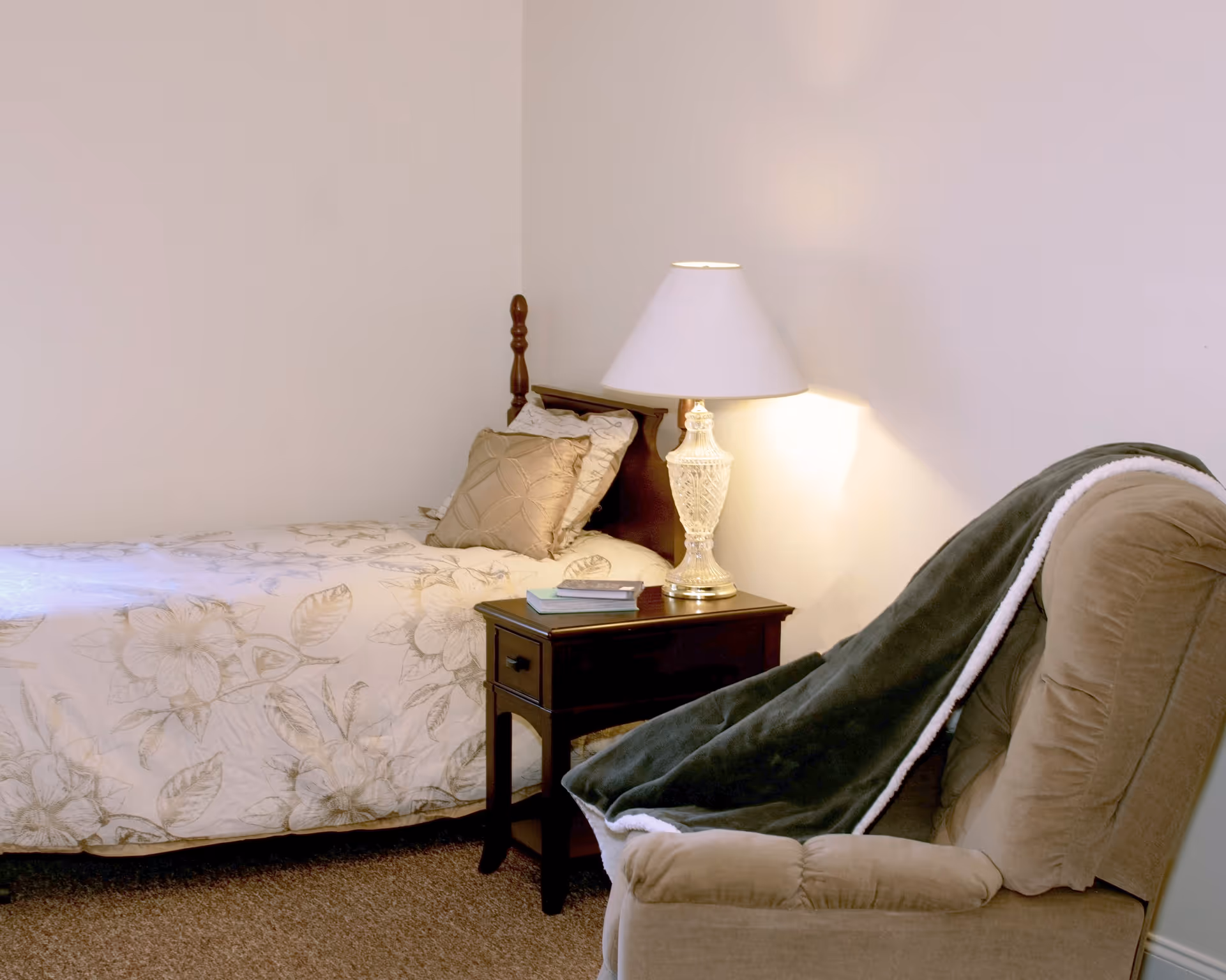 A cozy bedroom with a single bed covered in a floral patterned bedspread, a wooden nightstand with a decorative lamp and two books, and a comfortable upholstered armchair draped with a dark green blanket.