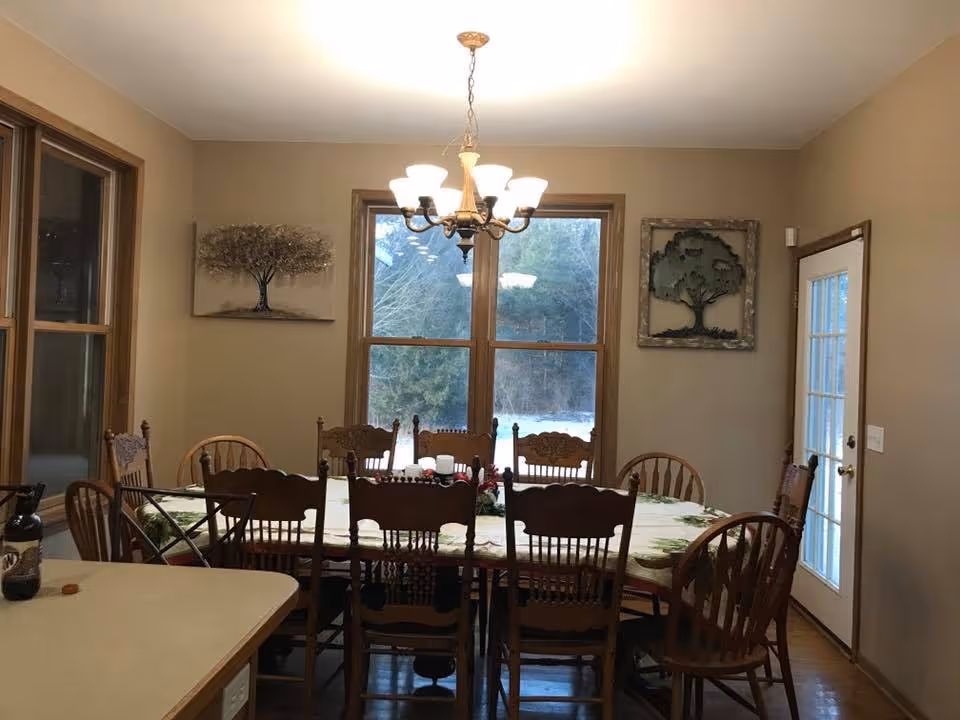 A dining room with a long wooden table covered with a tablecloth and surrounded by eight wooden chairs. There is a chandelier hanging above the table, two large windows showing an outdoor view with trees, and a glass door on the right side. The walls are beige with two tree-themed wall decorations.