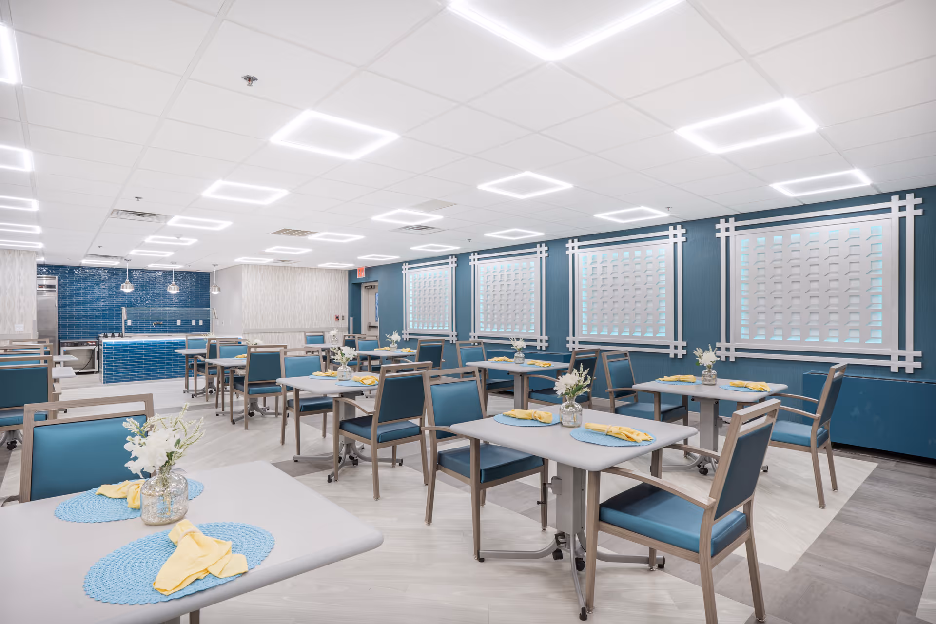 A bright, modern dining room with multiple tables and blue cushioned chairs. Each table is set with blue placemats, yellow napkins, and small flower arrangements in glass vases. The walls are painted blue with decorative white lattice panels, and the ceiling has square recessed lighting. In the background, there is a kitchen area with blue tiled walls and stainless steel appliances.