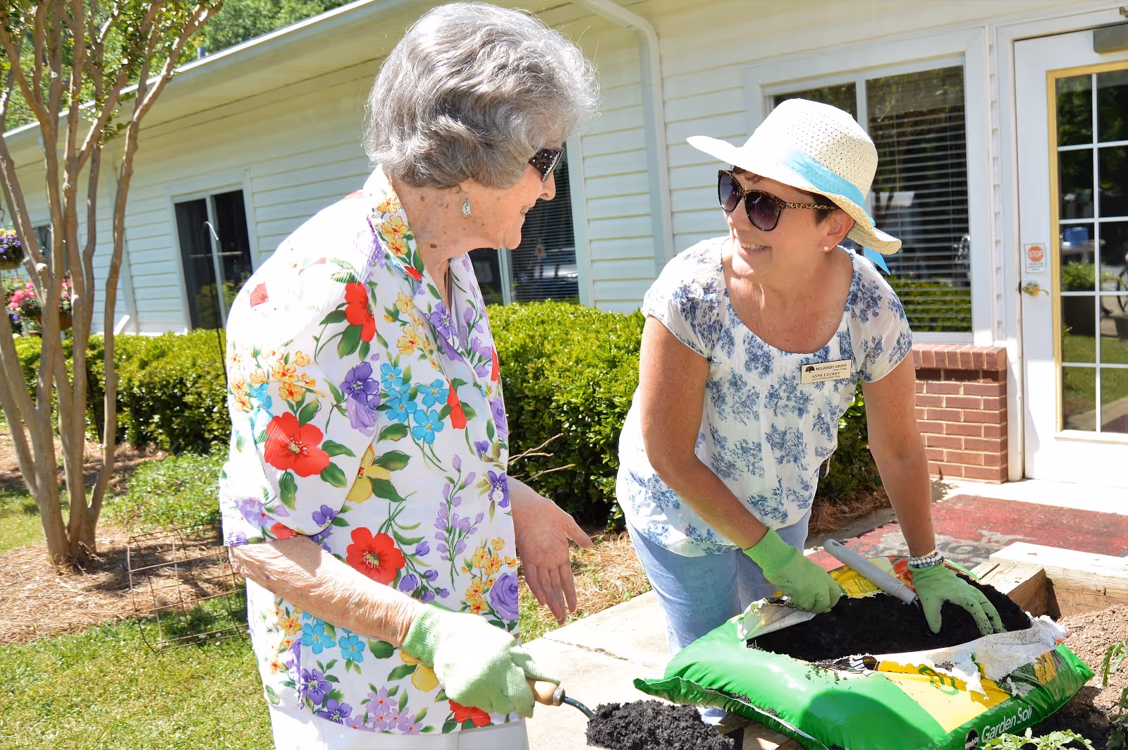 Two women gardening outside a building. One elderly woman wearing a floral shirt and green gloves is holding a small gardening tool, while the other woman wearing a hat, sunglasses, and a name tag is scooping soil from a bag labeled 'Garden Soil'. They are smiling and engaged in conversation near a garden bed with bushes and a tree in the background.