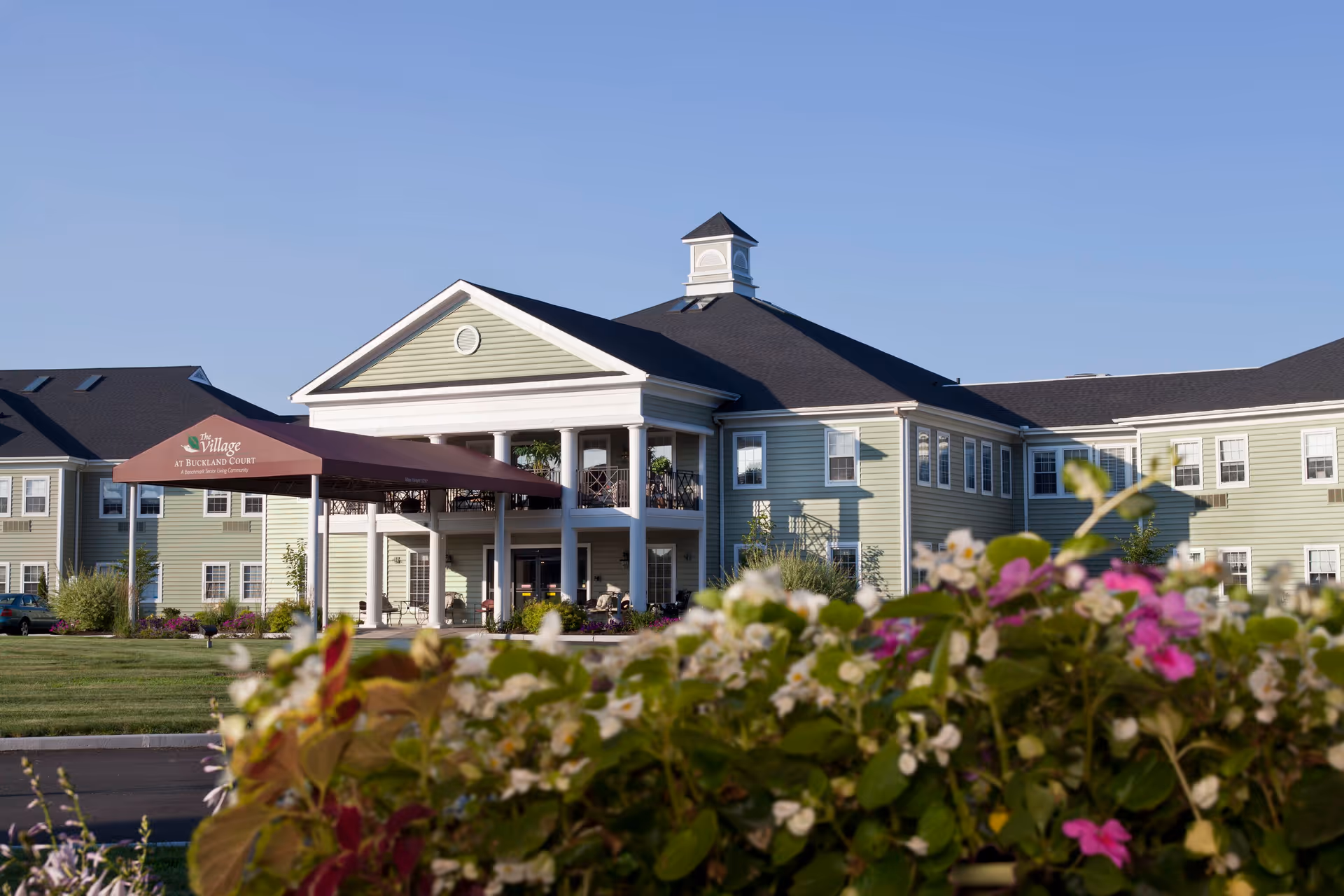 Exterior view of The Village at Buckland Court senior living facility with a green lawn and colorful flowers in the foreground under a clear blue sky.