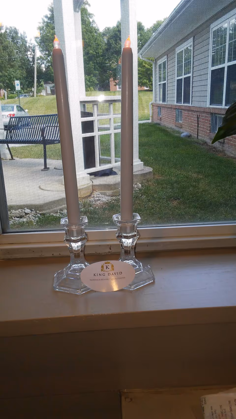 Two tall, lit candles in clear glass candle holders placed on a windowsill. Outside the window, there is a view of a grassy area, a bench, and part of a building with large windows.