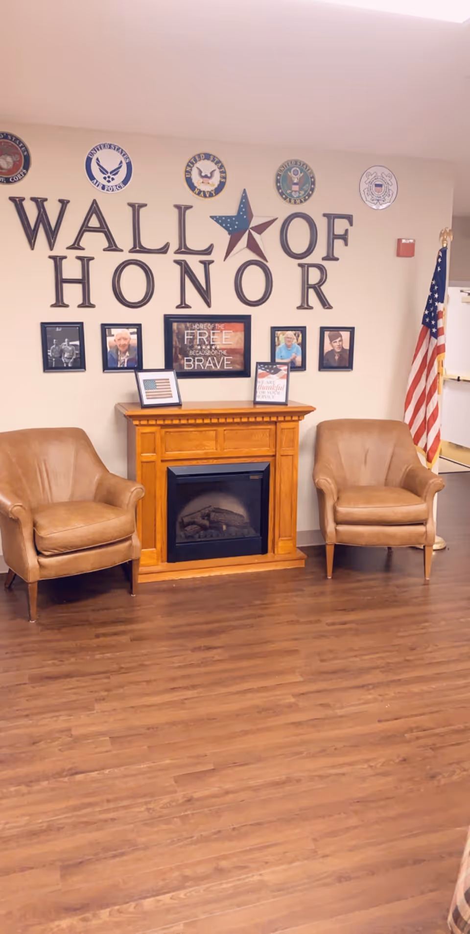 Indoor seating area with two leather chairs flanking a fireplace beneath a 'Wall of Honor' display and an American flag.