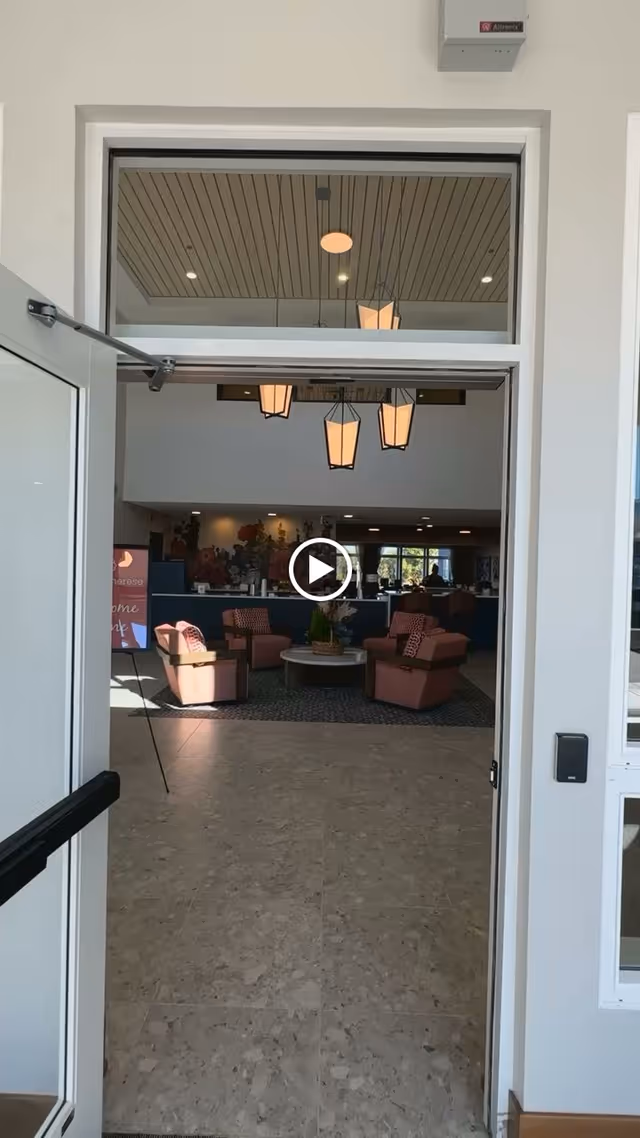 View through an open doorway into a senior living facility common area with four pink armchairs arranged around a round coffee table. The room has a high ceiling with hanging lantern-style lights and a mural on the far wall. There is a tiled floor and natural light coming from windows in the background.