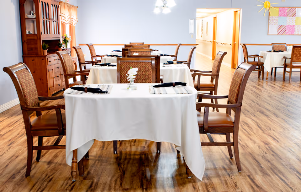 A dining room in a senior living facility with multiple tables covered in white tablecloths, each set with placemats, black napkins, and a small flower centerpiece. The room has wooden chairs with cushioned seats and backs, wood flooring, a wooden cabinet on the left side, and a hallway visible in the background.
