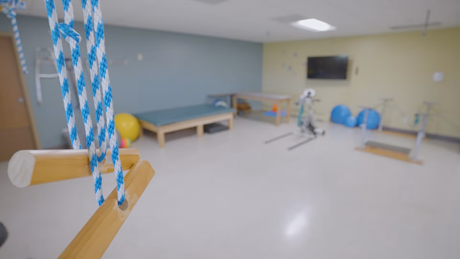 A physical therapy or exercise room with wooden trapeze bars hanging from blue and white ropes in the foreground. In the background, there are exercise mats, therapy tables, exercise balls, and various exercise equipment against pastel-colored walls with a mounted TV.