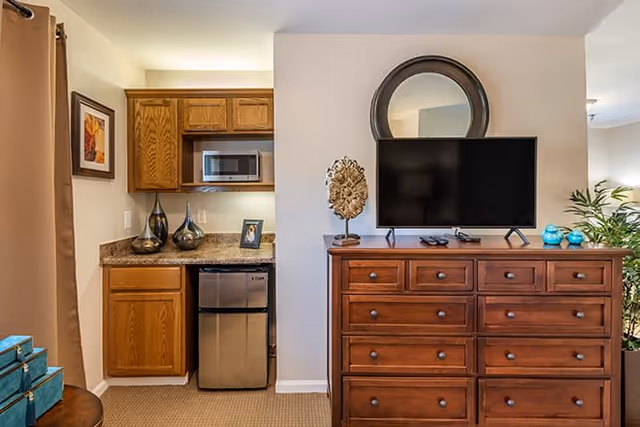 Interior view of a room featuring a wooden dresser with multiple drawers, a flat-screen TV on top, a round mirror mounted on the wall above the TV, and decorative items including a sculpture and blue vases. To the left, there is a small kitchenette area with wooden cabinets, a microwave, a mini refrigerator, and decorative vases on the countertop. The room has beige walls and carpeted flooring.