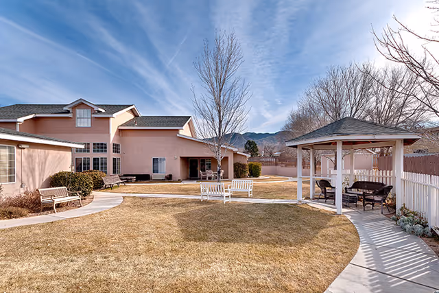 Outdoor courtyard area of a senior living facility with a gazebo, benches, and a pathway surrounded by grass and leafless trees. The building is light pink with multiple windows and a mountain range is visible in the background under a partly cloudy sky.