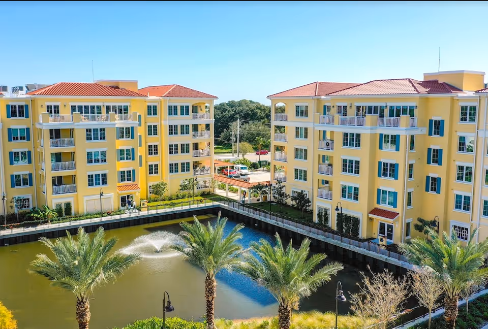 Exterior view of a senior living facility named Village On The Isle featuring two yellow multi-story buildings with red-tiled roofs surrounding a pond with a water fountain and palm trees.