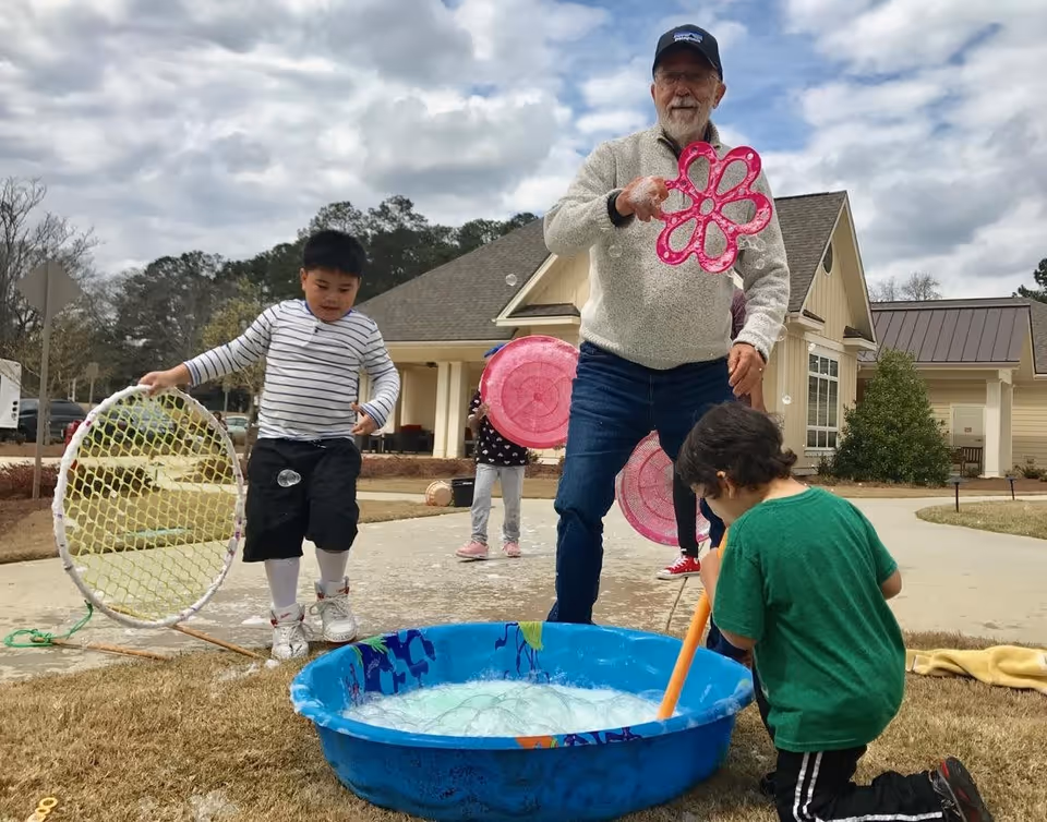 An elderly man and three children are playing outside near a senior living community building. The man is holding a large pink bubble wand, while one child in a striped shirt holds a large net, another child in a green shirt kneels near a blue plastic tub filled with bubble solution, and a third child stands in the background holding a pink bubble wand. The scene is set on a paved pathway with grass on either side and a cloudy sky overhead.