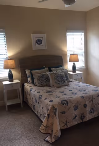 A bedroom with a bed covered in a seashell-patterned quilt and multiple decorative pillows. The bed has a woven headboard and is flanked by two white nightstands, each with a lamp. Two windows with blinds are on the wall behind the bed, and a framed picture hangs above the headboard.