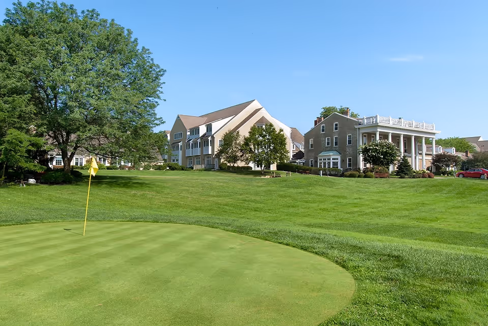 A well-maintained golf green with a yellow flag in the foreground, surrounded by lush green grass and trees. In the background, there are several large buildings with a clear blue sky above.