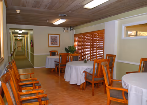 Interior view of a common area in Manatee River Assisted Living featuring round tables covered with white tablecloths and wooden chairs. The room has wooden flooring, a wooden ceiling, and a window with wooden blinds. A hallway extends from the room with handrails along the walls and framed artwork hanging.