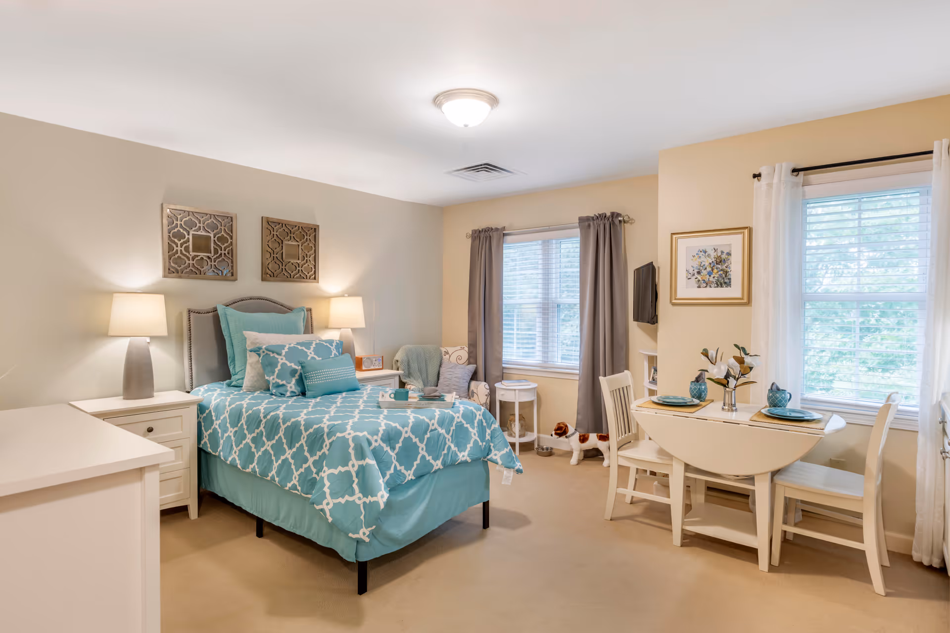 A cozy bedroom in an assisted living facility featuring a single bed with teal and white patterned bedding, two bedside tables with lamps, a small armchair with a throw blanket, a round side table, a drop-leaf dining table set with two chairs and place settings, two windows with curtains, and wall decorations including framed art and decorative mirrors.