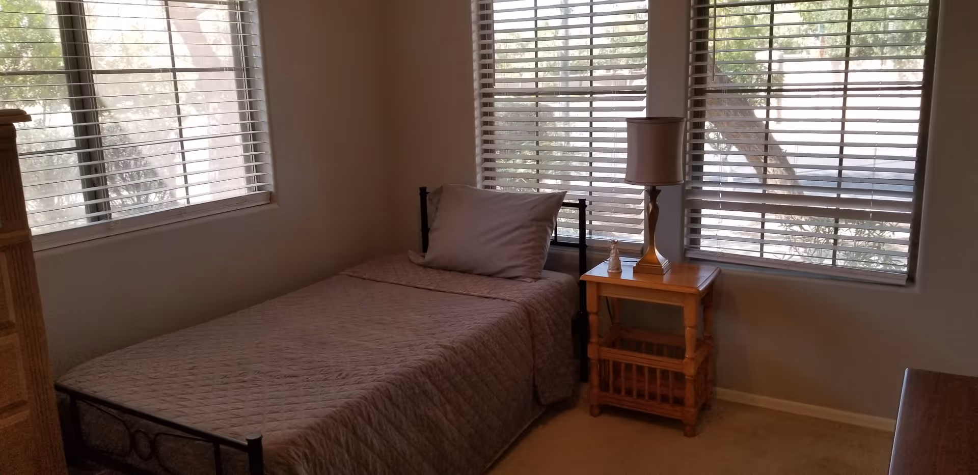 A small bedroom with a single bed covered in a gray quilt and a matching pillow. Next to the bed is a wooden nightstand with a lamp and a small decorative figurine. The room has two large windows with white blinds, allowing natural light to enter.