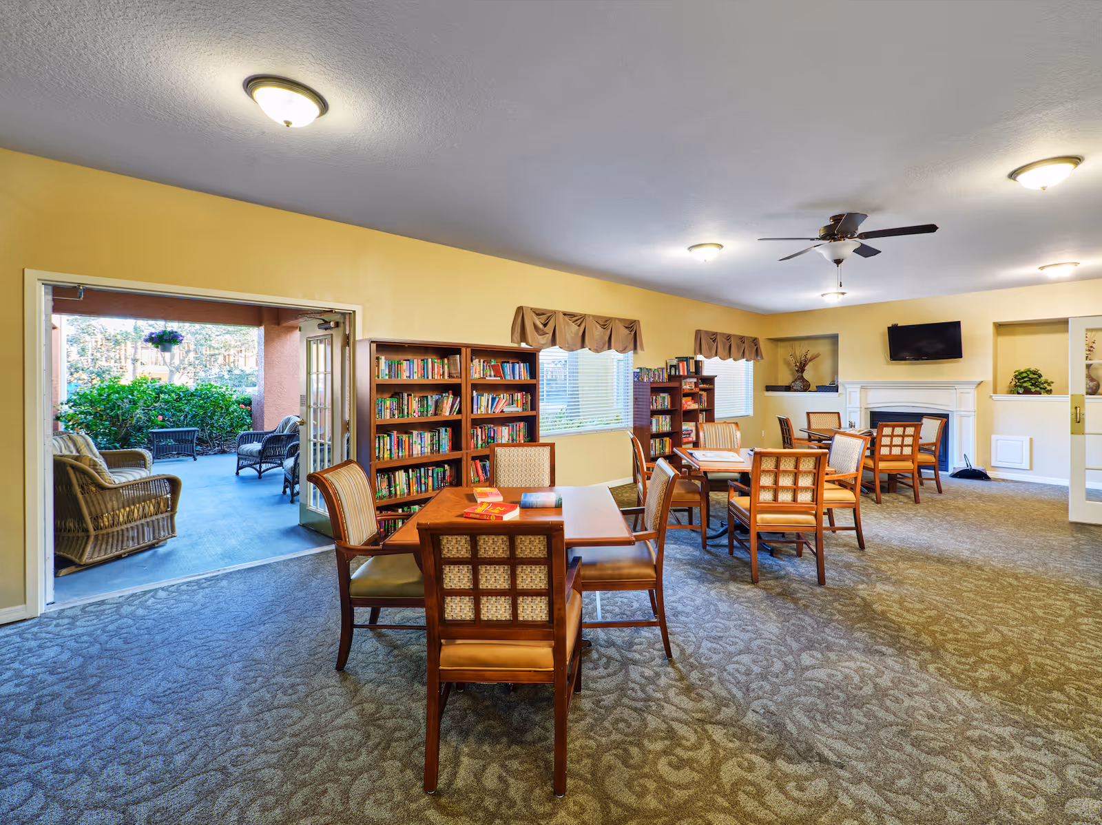 A cozy common area in an assisted living facility with several wooden tables and chairs arranged for socializing or activities. There are bookshelves filled with books along the yellow walls, windows with valances, a ceiling fan, and a wall-mounted TV above a white fireplace. Through an open door, an outdoor patio with wicker furniture and greenery is visible.