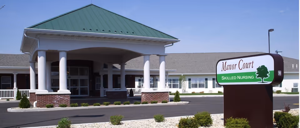 Exterior view of Manor Court Skilled Nursing facility with a covered entrance supported by white columns and a green roof. There is a sign in the foreground with the facility name and a tree logo. The building is single-story with multiple windows and a parking area in front.