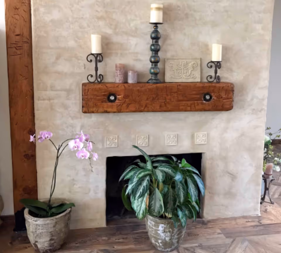 A cozy interior corner featuring a rustic fireplace with a wooden mantel. On the mantel are three candles on decorative holders and two small ornamental objects. In front of the fireplace are two potted plants, one with green leaves and the other with pink flowers. The wall has a textured finish and the floor is wooden.