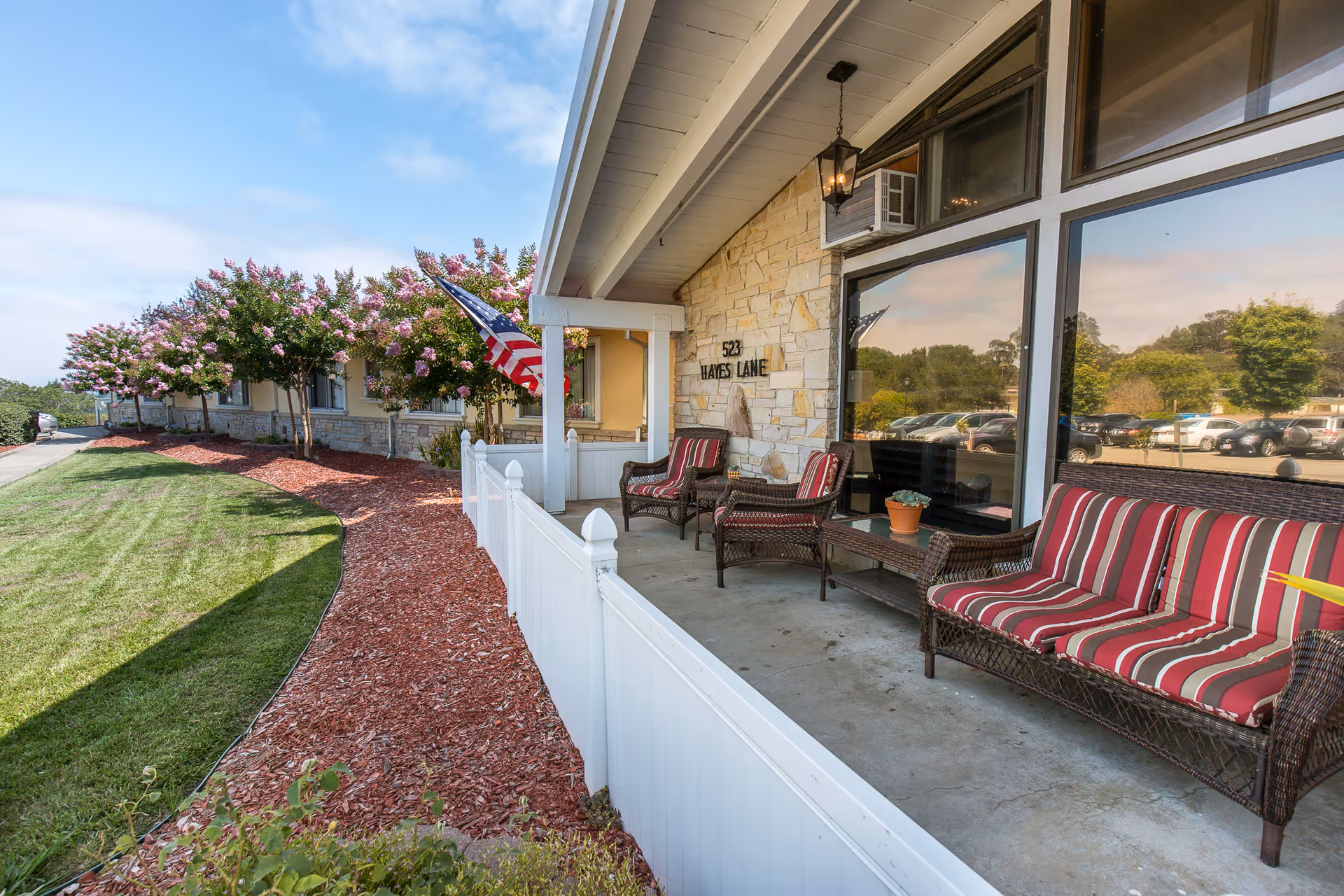 Covered front porch of a senior living facility with wicker seating and striped cushions, an American flag, and a landscaped lawn.