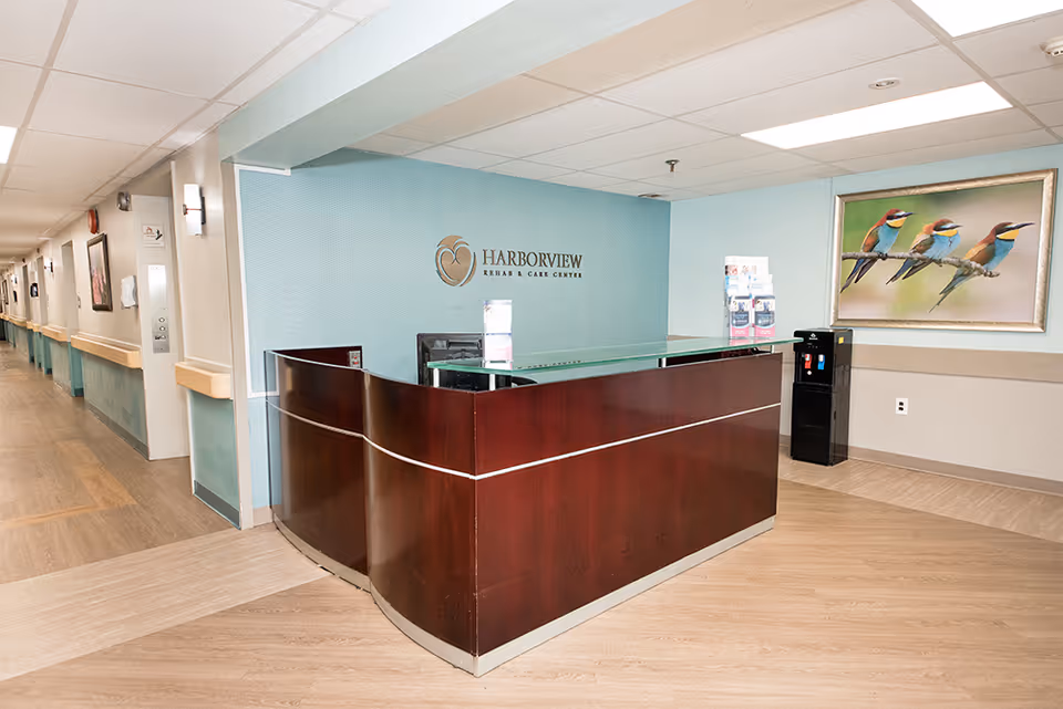 Reception area of Harborview Rehabilitation and Care Center at Doylestown with a curved wooden front desk, a blue accent wall behind it featuring the facility's logo, a water dispenser to the right, and a framed picture of colorful birds on the wall.