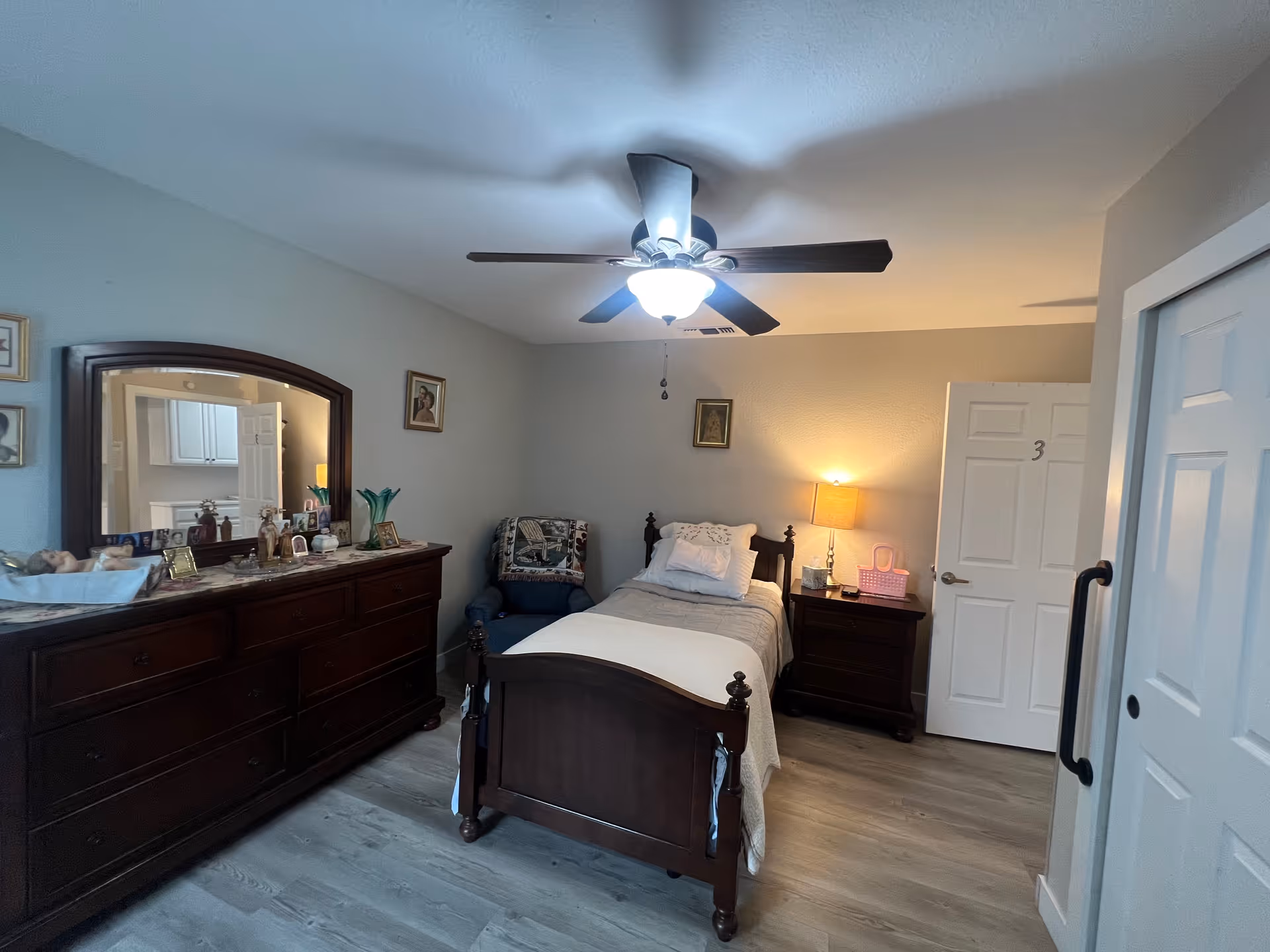 A cozy bedroom with a single wooden bed covered with white bedding, a wooden dresser with a large mirror, a blue armchair with a patterned blanket, a nightstand with a lamp and a pink basket, and a ceiling fan with lights. The room has light-colored walls and wood flooring.