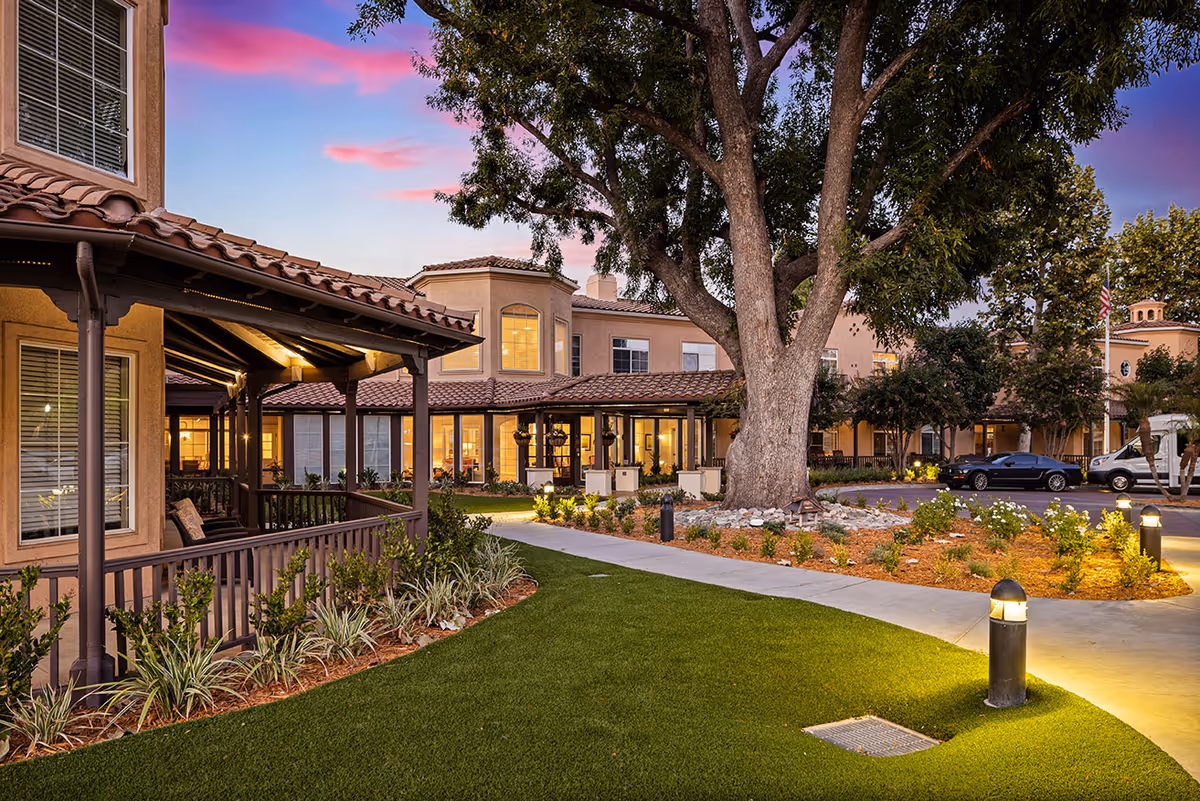 Exterior view of Ivy Park at West Hills senior living facility during sunset, showing a well-maintained garden with a large tree, a walkway, and a building with warm lights inside. There are parked cars and an American flag in the background.