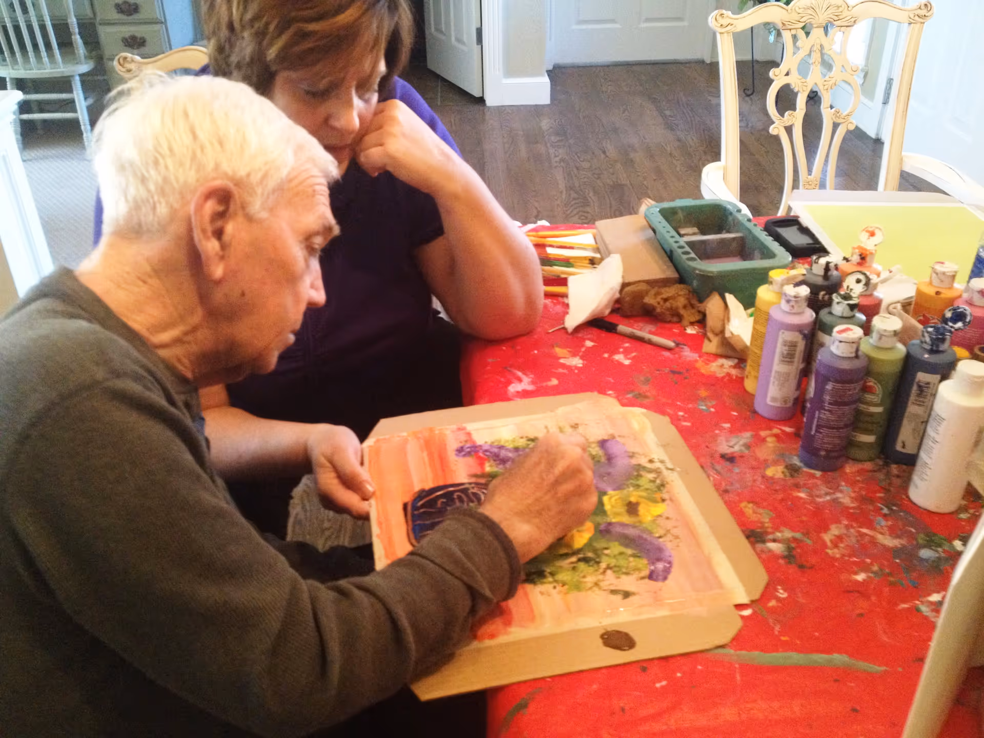 An elderly man painting on a piece of paper with various colors while a woman watches attentively. They are seated at a table covered with a red cloth, surrounded by bottles of paint and art supplies in a room with wooden flooring and white chairs.