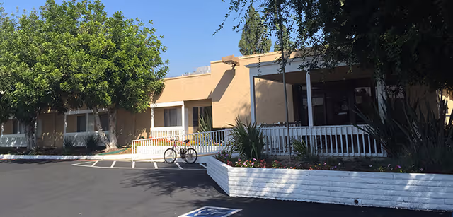 Exterior view of Monte Vista Healthcare Center showing a single-story beige building with a white railing, a wheelchair ramp, a bicycle parked near the entrance, and landscaped plants and trees under a clear blue sky.