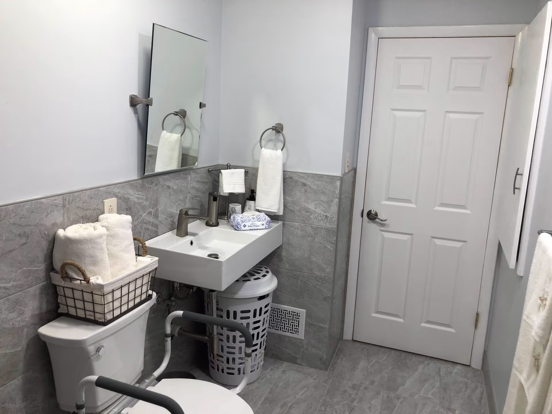 A clean and modern bathroom with gray tiled walls and floor. There is a white toilet with a basket of rolled white towels on top, a white sink with a silver faucet, a mirror above the sink, and a white door. A towel ring with a white hand towel is mounted on the wall next to the sink, and a white laundry basket is placed underneath the sink. The bathroom also features grab bars around the toilet for accessibility.