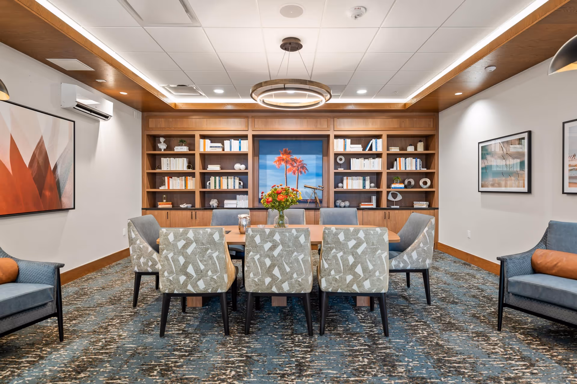 A dining room interior featuring a large table surrounded by patterned chairs, a bookshelf filled with books, and a vibrant painting of palm trees on the wall.