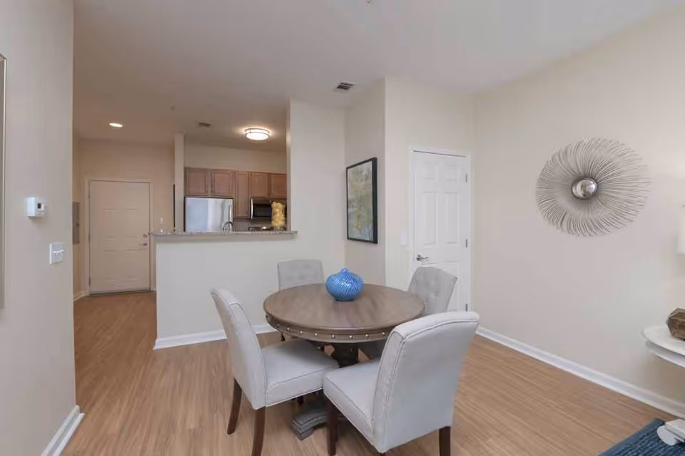 Interior view of a dining area with a round wooden table and four upholstered chairs. A blue decorative vase is placed on the table. The room has light-colored walls and wood flooring. In the background, there is a kitchen with wooden cabinets, a stainless steel refrigerator, and a microwave. A decorative wall piece and a framed picture are visible on the walls.