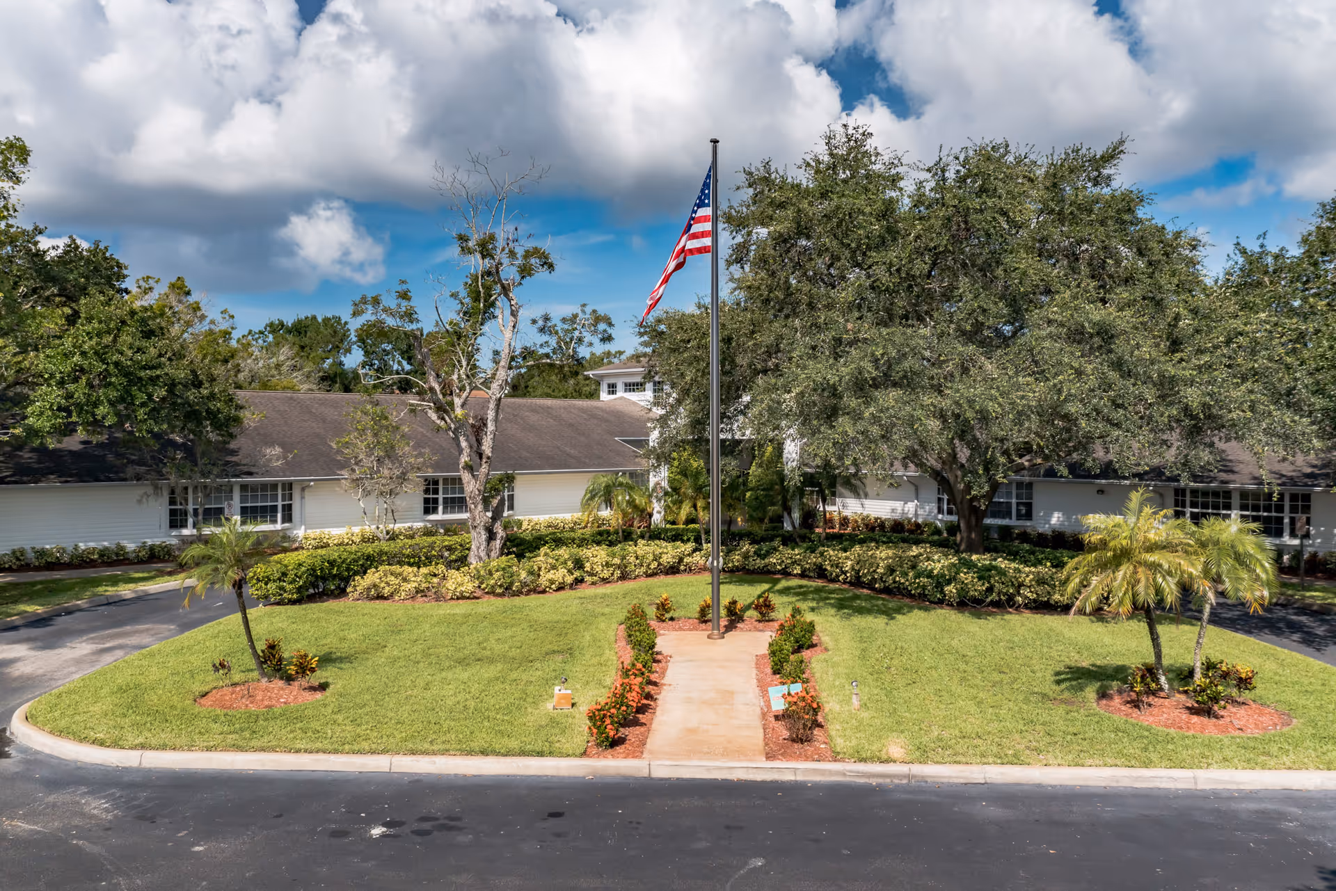 Front exterior view of a single-story building with a large green lawn, a flagpole with an American flag, and various trees and shrubs under a partly cloudy sky.