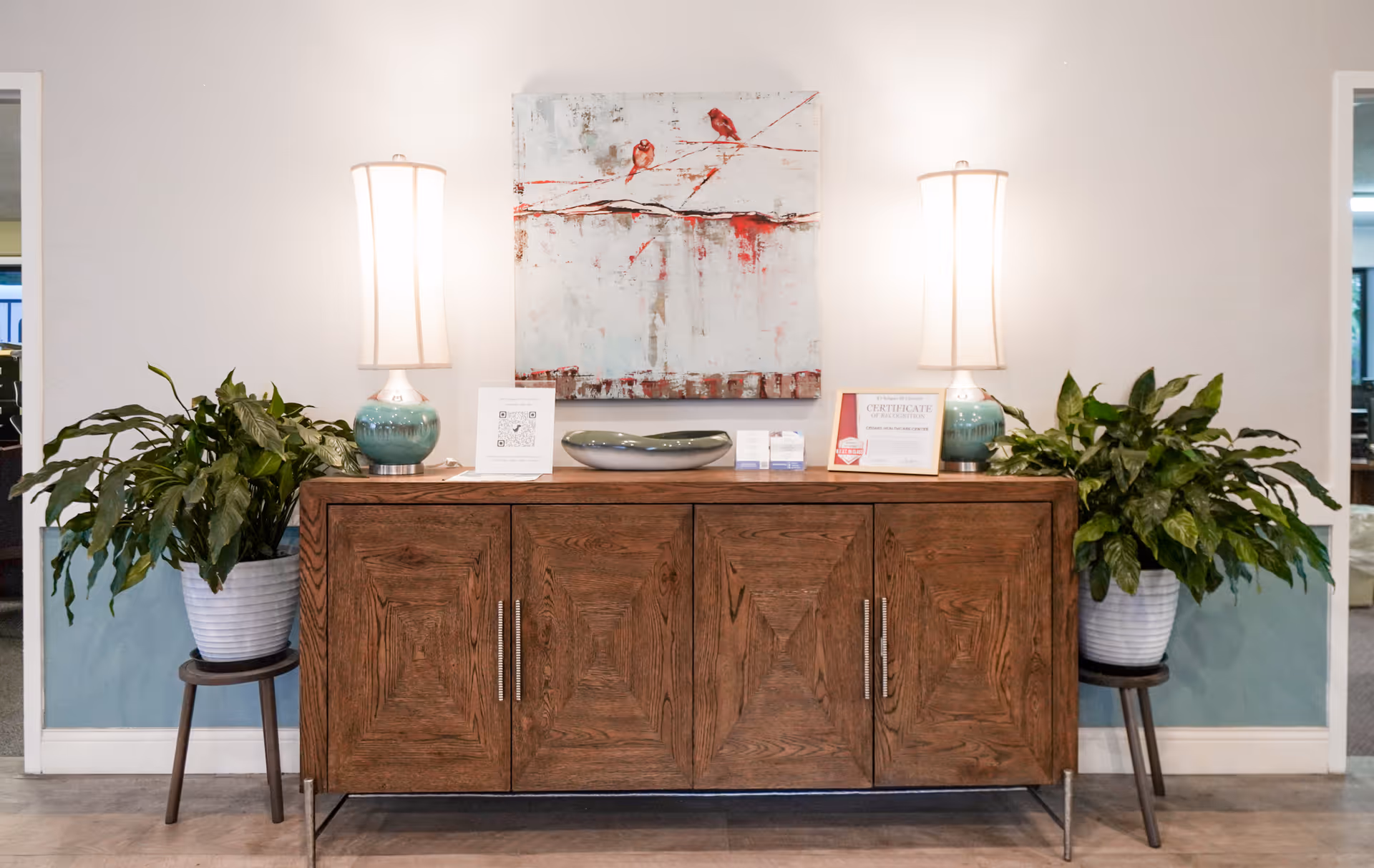 A wooden sideboard with four cabinet doors is positioned against a white wall. On top of the sideboard are two matching table lamps with teal bases and white shades, a decorative bowl, a certificate, and some small cards. Above the sideboard hangs a painting featuring birds on branches with red and white tones. On either side of the sideboard are green leafy plants in white pots placed on small wooden stands. The floor is light-colored wood, and the lower part of the wall is painted blue.