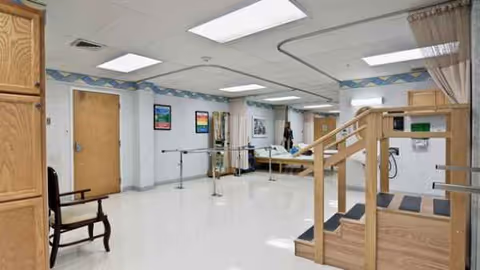 Interior rehab area with parallel bars, wooden staircase, chairs, and medical equipment.
