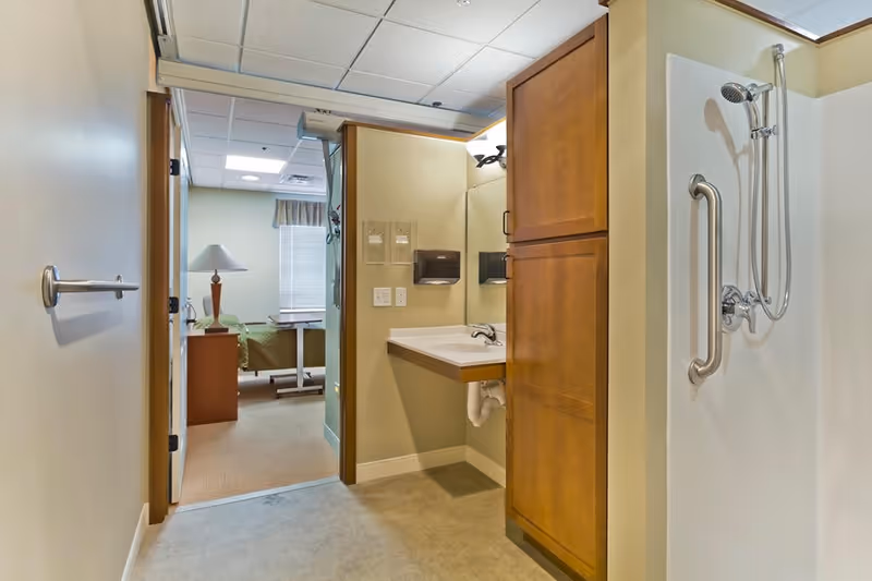 View of a senior living facility bathroom area with a wall-mounted sink, wooden cabinet, and a shower with a handheld showerhead and grab bar. In the background, a bedroom with a bed, bedside table, lamp, and window with blinds is visible.