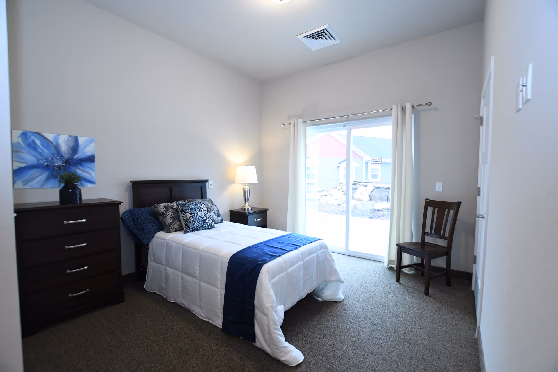 Bright furnished bedroom featuring a single bed with blue accents, a dresser, nightstand with lamp, chair, and sliding glass doors to the outside.