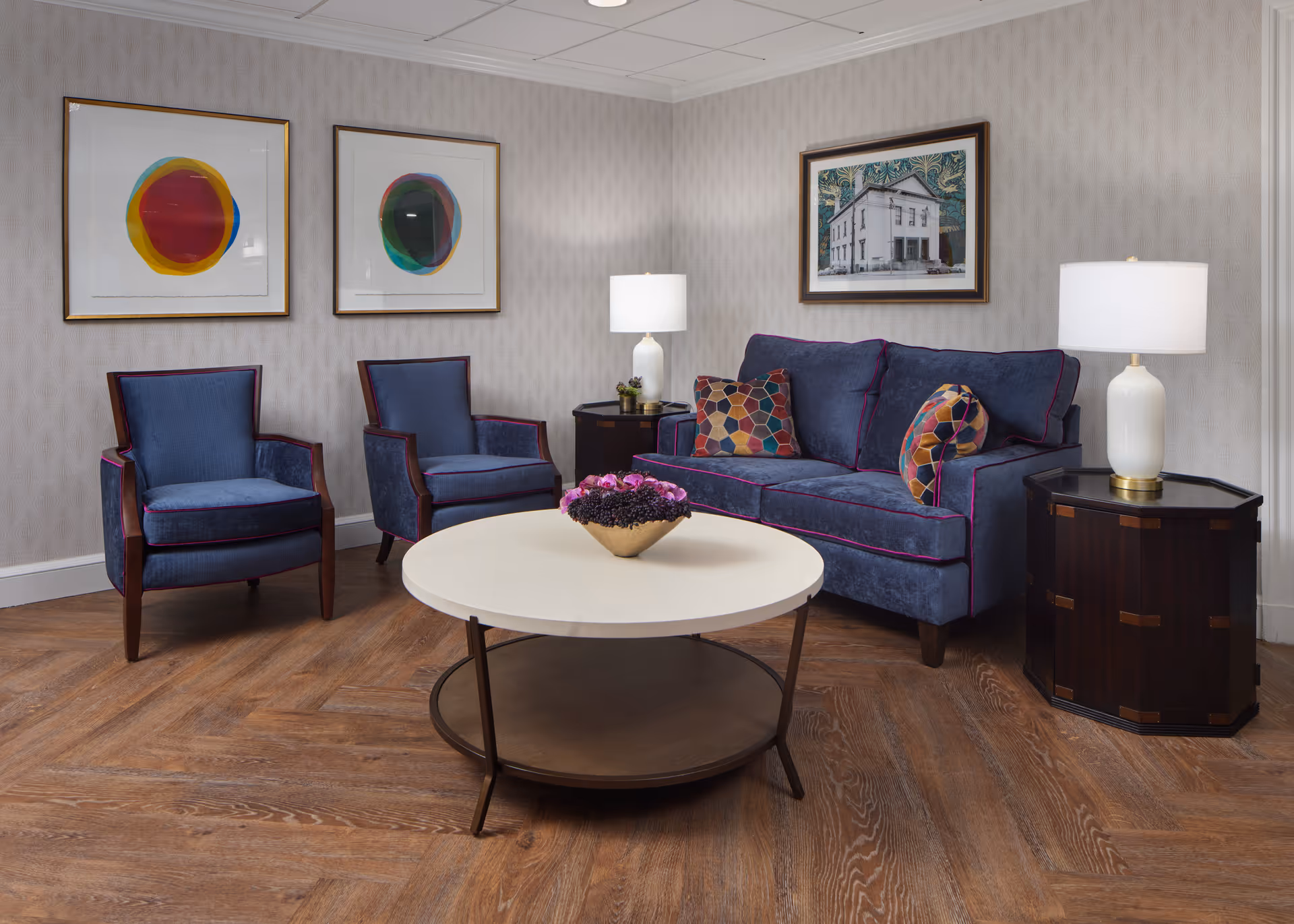 A cozy living room area with a blue sofa adorned with colorful patterned pillows, two matching blue armchairs, a round white coffee table with a flower arrangement, two white table lamps on dark wooden side tables, and three framed artworks on the light gray patterned walls.