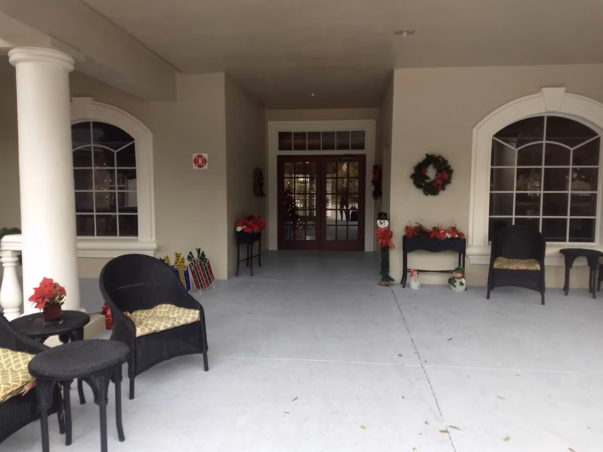 Covered entrance area of a building with two large arched windows, black wicker chairs with cushions, small tables, and holiday decorations including wreaths, poinsettias, and snowman figures near the double glass doors.