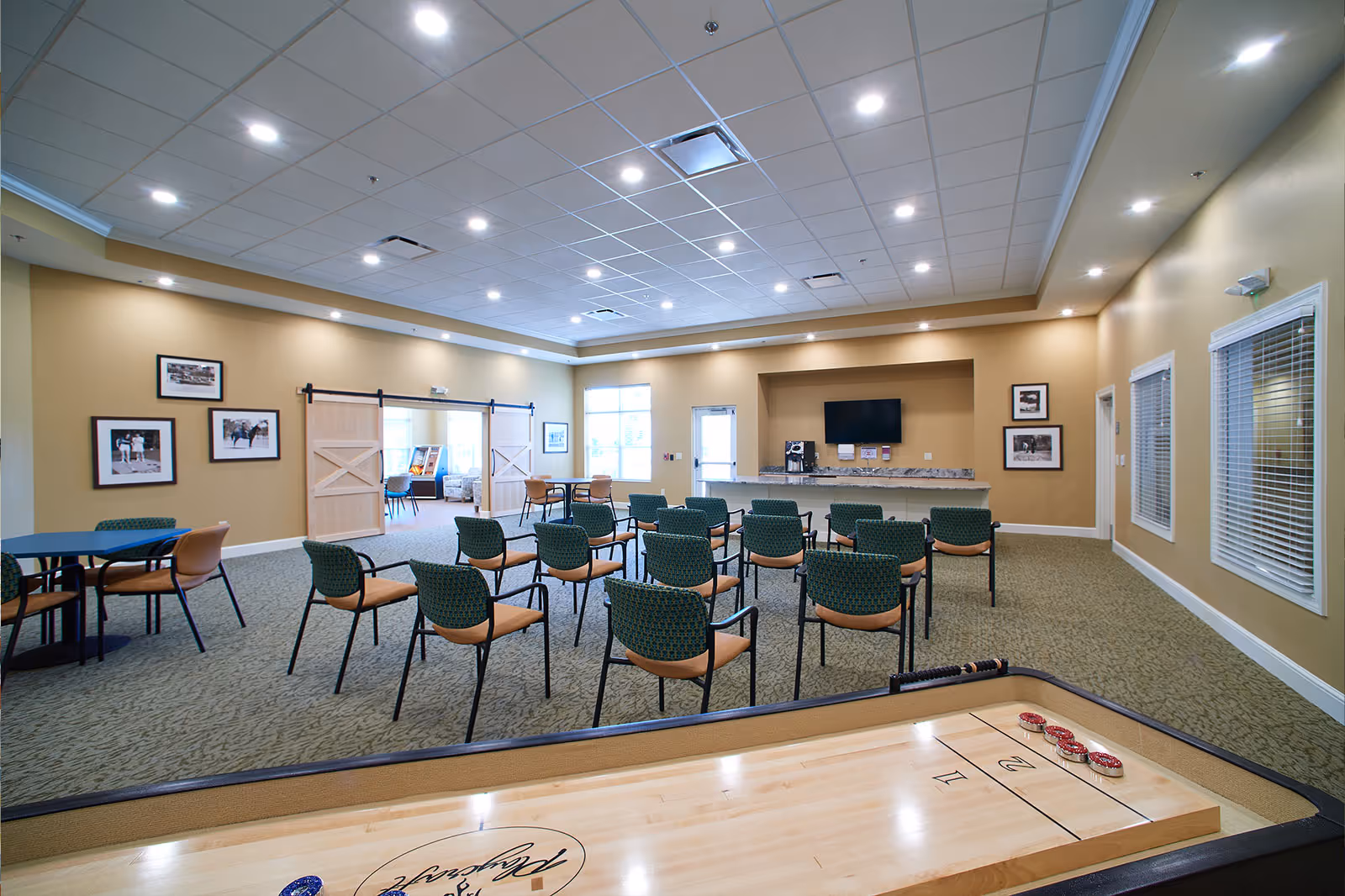 Spacious interior activity room with rows of chairs facing a TV and counter, tables to the side, and a shuffleboard in the foreground.