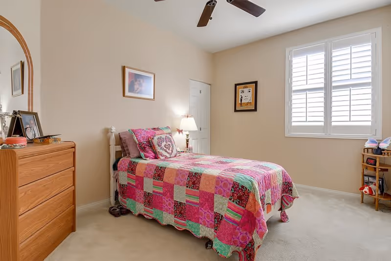 A cozy bedroom with a single bed covered in a colorful patchwork quilt in shades of pink, green, and red. The room has beige walls, a ceiling fan, a wooden dresser with a mirror, framed pictures on the walls, a window with white shutters, and a small wooden shelf with toys and books.