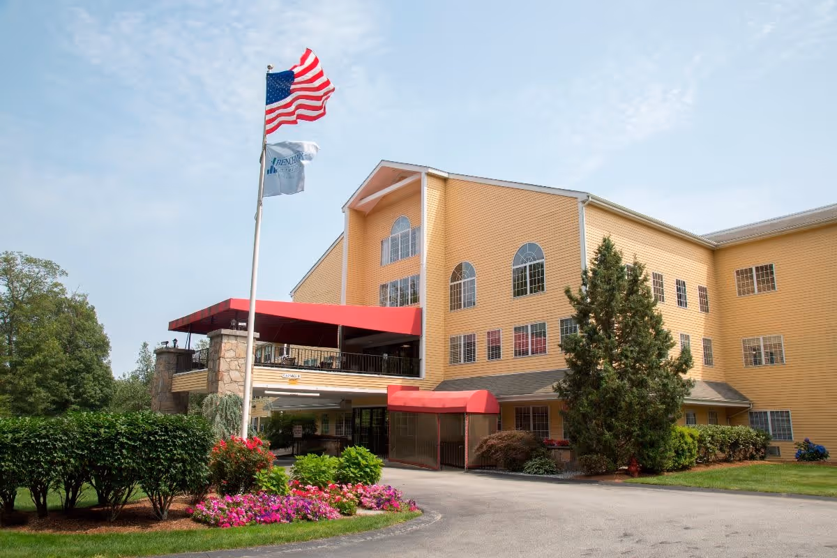 Exterior view of a multi-story yellow building with large windows and a red awning over the entrance. There are two flagpoles in front, one with an American flag and the other with a white flag. The surrounding area includes green bushes, colorful flowers, and a paved driveway.